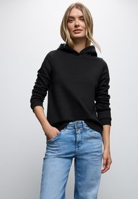 Felpa nera con cappuccio e polsini a costine, abbinata a jeans di denim azzurro chiaro con chiusura a bottoni e tasche. Design minimalista.