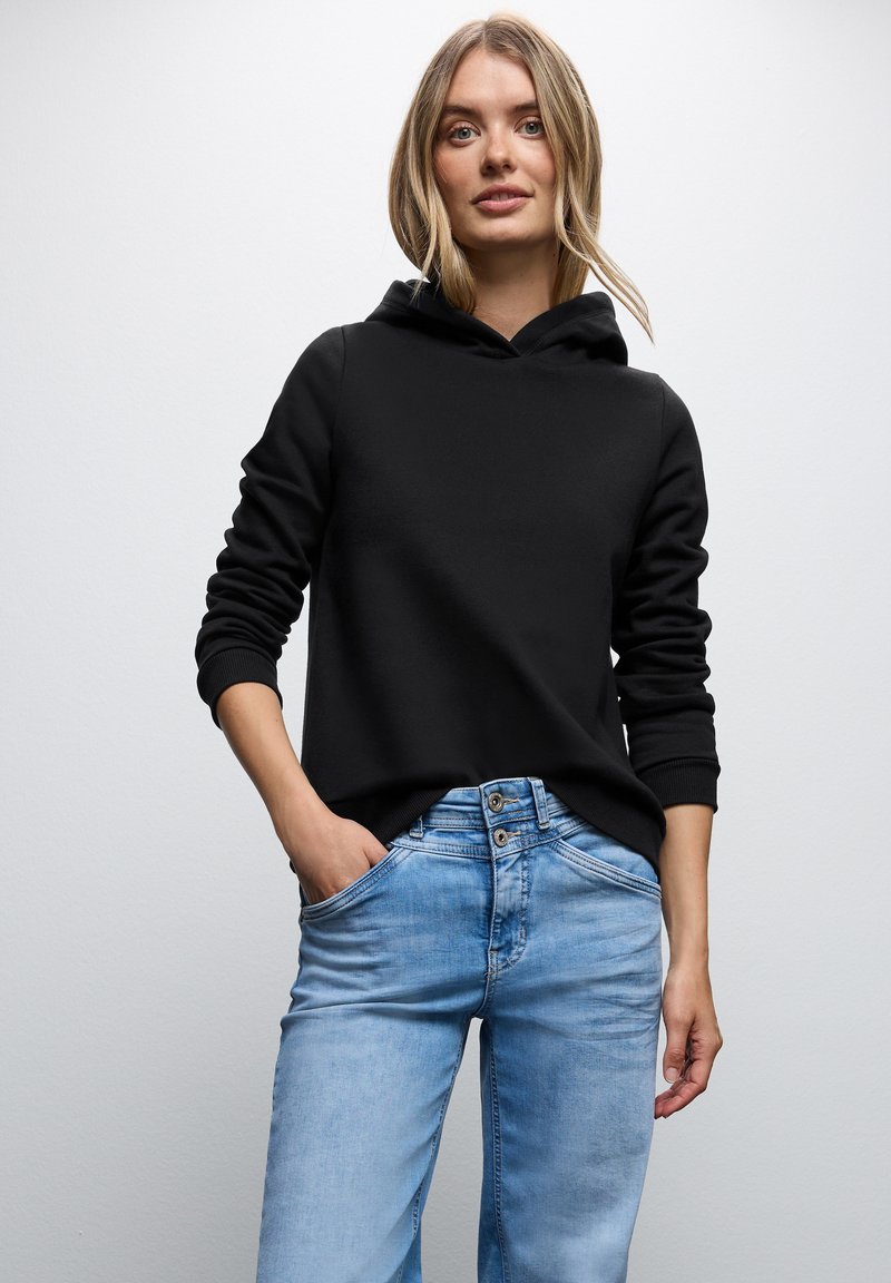 Felpa nera con cappuccio e polsini a costine, abbinata a jeans di denim azzurro chiaro con chiusura a bottoni e tasche. Design minimalista.