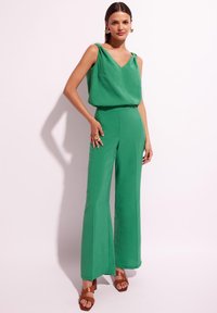 Zielony, bezrękawnikowy kombinezon z dekoltem w szpic, zwiewny materiał, szerokie nogawki, podkreślona talia, połączony z brązowymi sandałami na obcasie.