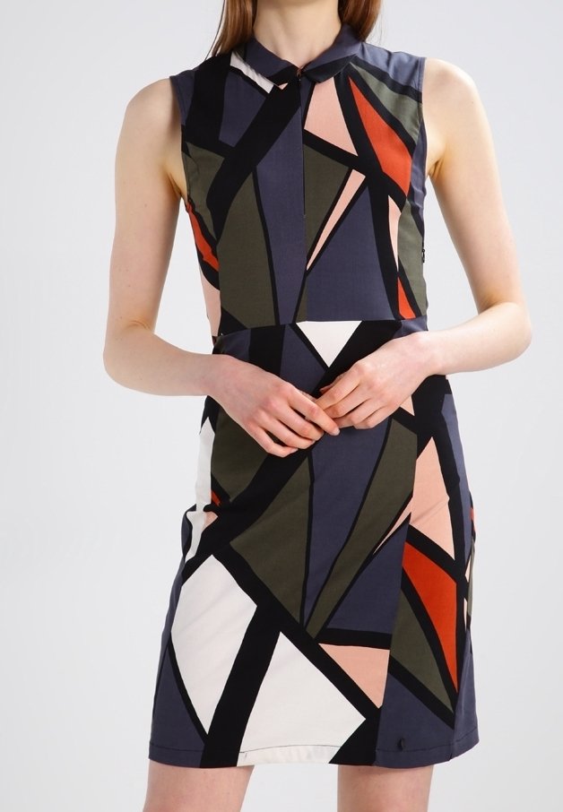 Ärmlös klänning med geometriskt mönster i svart, olivgrönt, marinblått, rött, vitt och rosa, bärs av en person med händerna knäppta vid midjan.