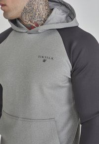 Felpa grigia e grigio scuro con tasca frontale, caratterizzata da un logo sul petto. Il materiale sembra liscio con un mix di texture.