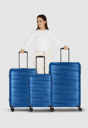 Femme souriante tenant les poignées de trois valises rigides bleues de taille petite, moyenne et grande, chacune dotée de quatre roues.