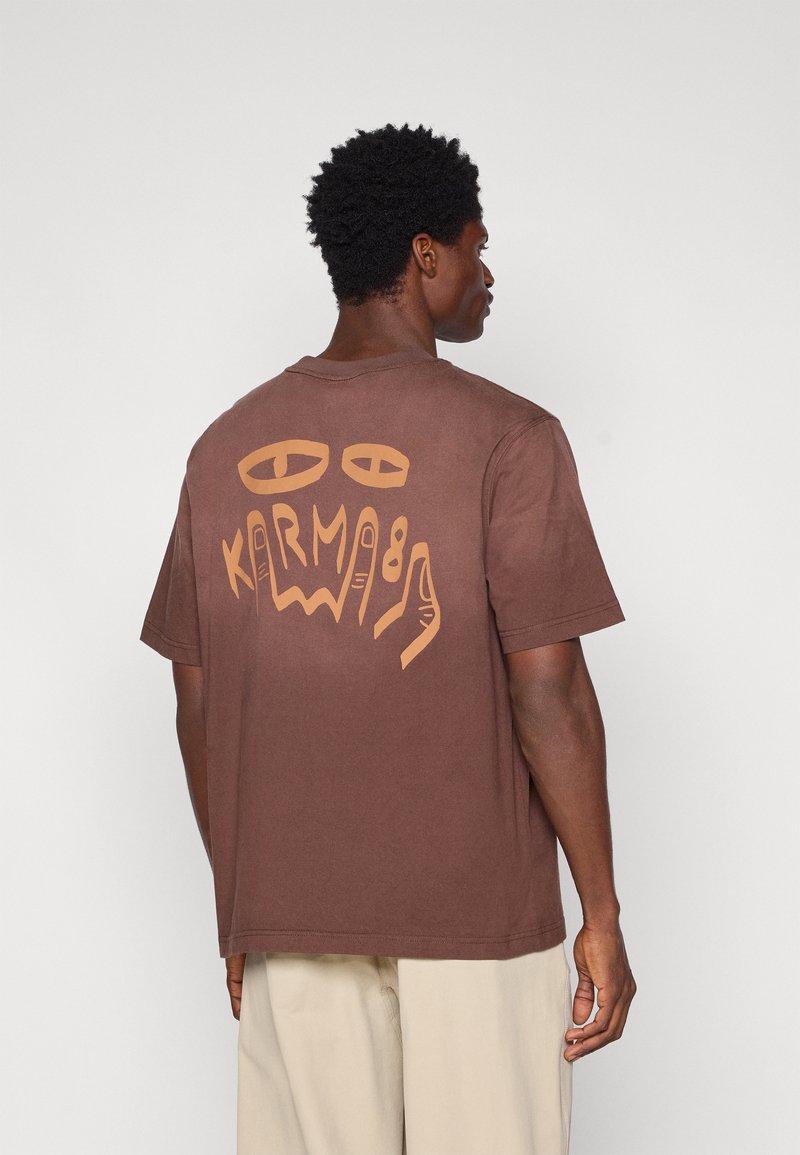 T-shirt in cotone marrone con una grande grafica astratta color crema sulla schiena, caratterizzata da occhi e dita stilizzati. Maniche corte, vestibilità rilassata.