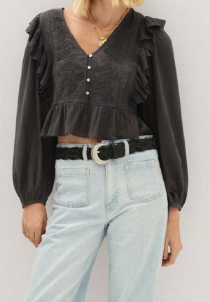 Kobieta ubrana w czarną, długorękawową bluzkę z falbankami i guzikami oraz jasnoniebieskie, wysokowalne dżinsy z czarnym plecionym paskiem.
