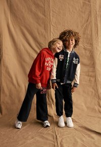 Zwei Kinder stehen zusammen vor einem beigen Hintergrund. Eines trägt einen roten Kapuzenpulli und marineblaue Hosen, während das andere eine marineblaue Collegejacke mit weißen Ärmeln und schwarze Hosen trägt.