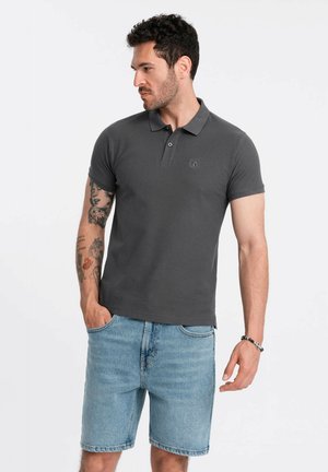 Dunkelgraues Poloshirt mit kurzen Ärmeln, ausgestattet mit einem Kragen und einem Logodetail. Kombiniert mit hellblauen Denim-Shorts, steht das Model mit den Händen in den Taschen.