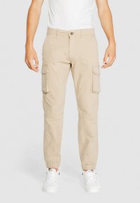 Beige Cargo-Hosen mit elastischen Bündchen, mehreren Seitentaschen und einer normalen Taille. Getragen mit einem weißen T-Shirt und weißen Sneakers.