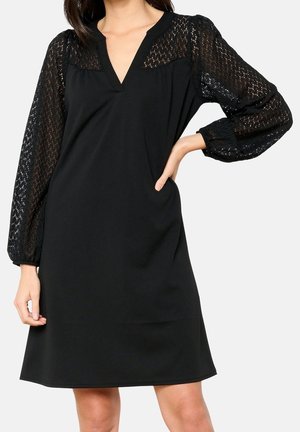 Czarna sukienka do kolan z długimi, przezroczystymi koronkowymi rękawami i dekoltem w kształcie litery V, noszona przez kobietę z jedną ręką na biodrze.