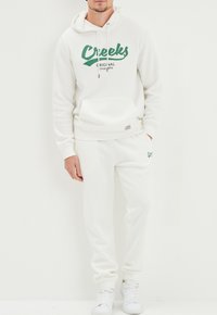 Felpa bianca con testo verde "Creeks Original dal 1974" e tasca frontale. Abbinata a pantaloni jogger bianchi e sneakers bianchi.