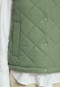 Gilet vert matelassé avec des motifs en losange, poche latérale et détails de fermeture à pression. Superposé sur une chemise blanche. Texture lisse.