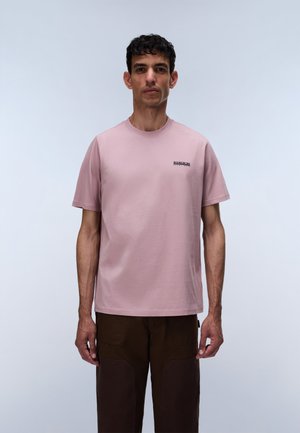 Homme aux cheveux courts et foncés portant un t-shirt rose clair à manches courtes et un pantalon marron foncé, debout devant un fond clair uni.