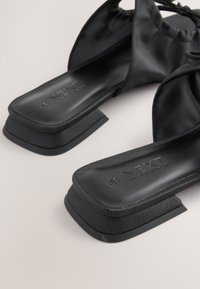 Schwarze Slide-Sandalen mit strukturiertem Material, eckigem Absatz und einer glatten Innensohle mit eingeprägtem Branding. Das Design ist minimalistisch.