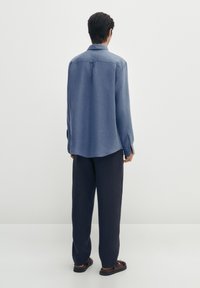 Blå skjorta gjord av lättviktsmaterial med avslappnad passform, med långa ärmar, en spetsig krage och en rundad nederkant. Bärs med mörka byxor.