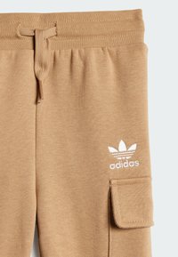 Gaišbrūnas sporta bikses no mīksta auduma ar regulējamu jostasvietu, sānu kabatu un baltu Adidas logu akcentu.