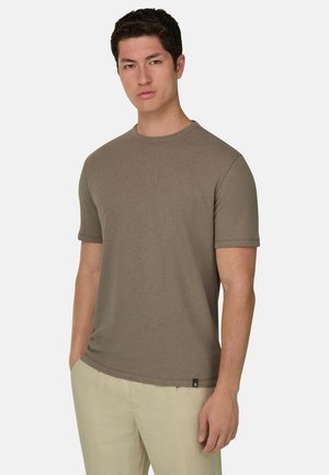 Homme portant un t-shirt olive à manches courtes et col rond, ainsi qu'un pantalon beige clair, debout avec les mains partiellement dans les poches, sur un fond uni.