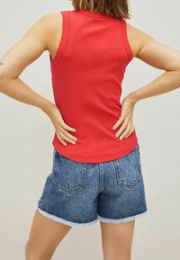 Red, ribbed tank top paired with blue denim shorts featuring frayed hems and back pockets. The model stands with hands on hips.