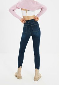 Jeans de mezclilla oscuros y ajustados con bolsillos traseros, complementados con una camiseta corta blanca y botas beige hasta el tobillo con suelas gruesas.