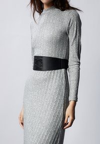 Ceinture noire large avec des détails lacés portée sur une robe grise à côtes. La robe présente des manches longues et une silhouette ajustée.