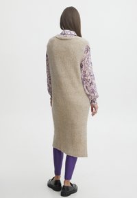 Gilet di maglia a coste beige con fessure laterali, indossato sopra una camicia a maniche lunghe floreale viola, abbinato a leggings viola e scarpe nere.