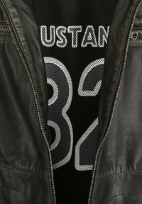 Schwarze Lederjacke mit Reißverschluss, die ein schwarzes Netzshirt enthüllt, das weißen Text "USTAN" und die Zahl "32" in einem fetten, durchbrochenen Design zeigt.