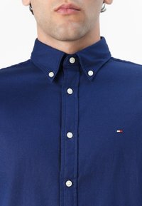 Camisa de botones azul marino con un collar clásico, que presenta botones blancos y un pequeño logo con acentos en rojo y blanco en el lado izquierdo.