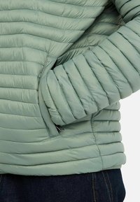 Lys grøn pufferjakke med quiltet tekstur, der har vandret syninger, sidelommer og en glat overflade.
