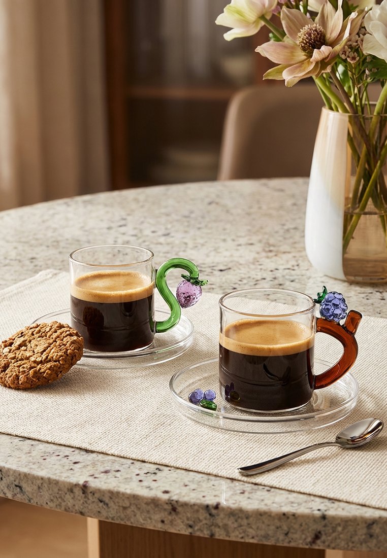 Dwie filiżanki do espresso z dekoracyjnymi uchwytami w kształcie owoców na przezroczystych spodkach, ustawione na beżowym bieżniku, obok znajduje się ciastko owsiane i łyżeczka.