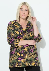 Blondýnka v multicolor kamuflážovém topu s třičtvrtinovými rukávy, dotýkající se svých vlasů, stojící o bílou zeď.