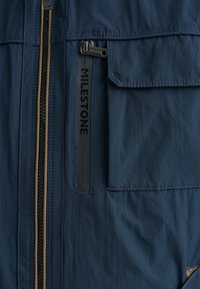 Marineblaue Jacke mit schwarzem Reißverschluss, ausgestattet mit einer großen Brusttasche und strukturiertem Stoff. Markenkennzeichnung "MILESTONE" am Reißverschlussanhänger.