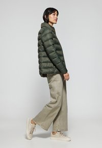 Olivgrön pufferjacka med hög krage, quiltad yta och åtsittande passform. Kombinerad med vida beiga byxor och ljusa sneakers.