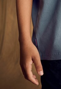 Světle modré bavlněné tričko na dětském paži, s červenou etiketou "Superdry" na boku, hladká textura, krátké rukávy.