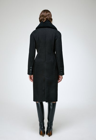 Femme aux cheveux bouclés portant un long manteau noir avec un large col et des bottes noires à talons hauts jusqu'aux genoux, debout de dos.