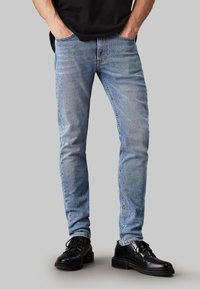 Jasnoniebieskie wąskie dżinsy z przetartym praniem, z pięcioma kieszeniami, pasami na pasek i przetarciami na kolanach. Zestawione z czarnymi butami.