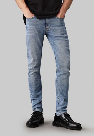 Jeans aderenti blu chiaro con una finitura lavata, featuring cinque tasche, passanti per cintura e dettagli strappati sulle ginocchia. Abbinati a scarpe nere.