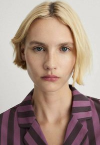 Košeľa s krátkym rukávom v tmavofialových a burgundských pruhoch, s výstrihom typu notch. Modelka má krátke blond vlasy a hladkú pleť s pehami.