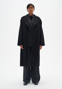 Manteau en laine noir avec un large col, une silhouette oversize et des manches longues, porté sur une chemise noire et un pantalon large. Texture lisse.
