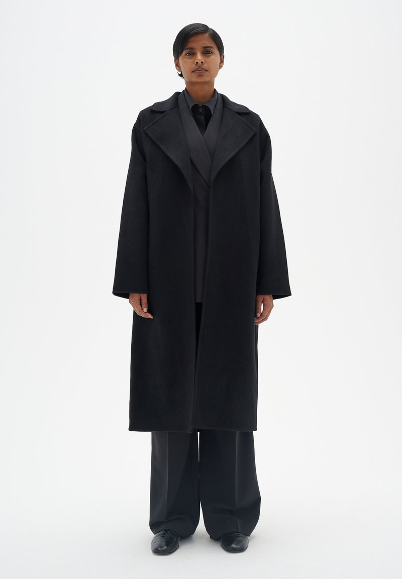 Manteau en laine noir avec un large col, une silhouette oversize et des manches longues, porté sur une chemise noire et un pantalon large. Texture lisse.