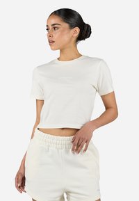 Tricou scurt la jumătate cu mânecă scurtă din bumbac crem, având un logo reflectorizant pe piept. Asortat cu șorturi crem corespunzătoare.
