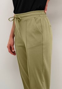 Svetlo olivno zelene hlače z sproščenim krojem, elastičnim pasom in vrvico. Imajo stranske žepe in gladko, rahlo teksturirano tkanino.