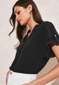Blusa nera a maniche corte con scollo a V e dettagli plissettati sulle spalle, abbinata a pantaloni bianchi a vita alta. Tessuto dalla texture liscia.