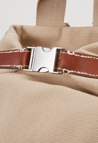 Gros plan d'un sac en tissu beige avec une bandoulière en cuir marron et une boucle en métal argenté fixant la sangle.