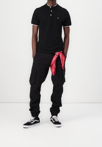 Man in een zwart polo met korte mouwen, zwarte cargo joggers met elastische boorden, zwart-witte sneakers en een rode bandana die uit zijn zak hangt.