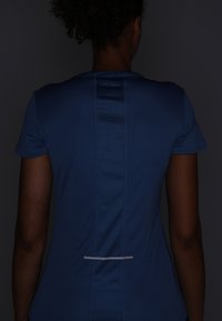 Maglietta sportiva blu con maniche corte, pannello in rete testurizzato lungo il centro della schiena e dettaglio riflettente vicino all'orlo.