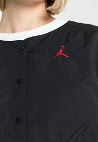 Svart jacka med en texturerad fabrik, prydd med en vit krage och svarta knappar. Anmärkningsvärt röd Air Jordan-logotyp på bröstet.