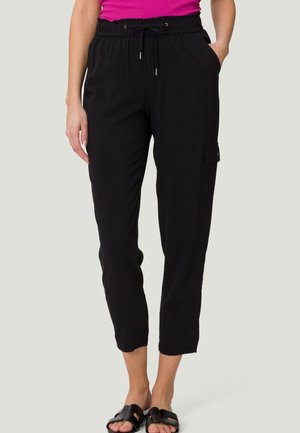 Frau trägt schwarze Cargo-Hose mit Kordelzug in der Taille und Seitentasche, kombiniert mit schwarzen Slidesandalen und einem pinkfarbenen ärmellosen Top.