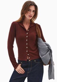 Bordeaux knap cardigan med hætte, prydet med ribbet tekstur og sølvknapper, parret med mørke jeans og holder en grå sweater.