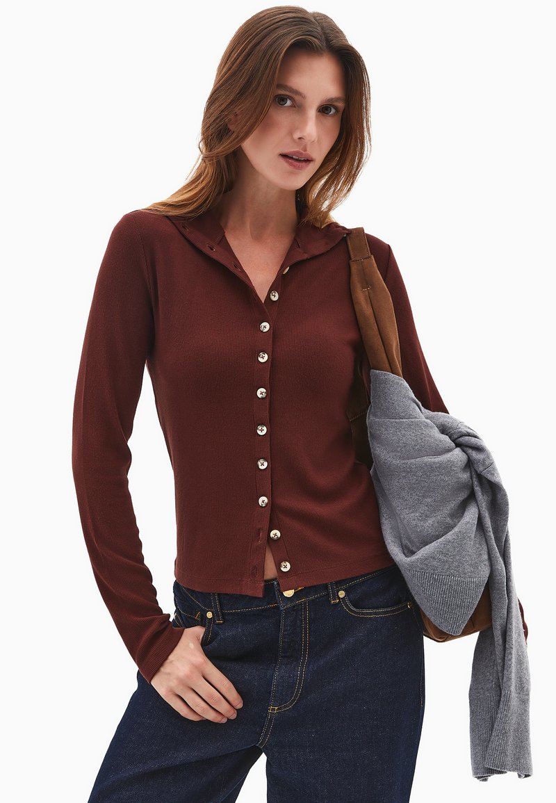 Bordeaux knap cardigan med hætte, prydet med ribbet tekstur og sølvknapper, parret med mørke jeans og holder en grå sweater.