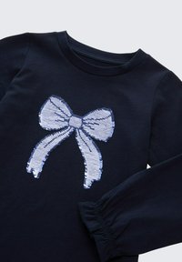 Navyblaues Langarmshirt mit einer großen, mit Pailletten versehenen, hellblauen Schleifen-Grafik in der Mitte. Die Ärmel haben ein gerafftes Manschettendetail.