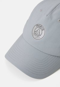 Lichtgrijze honkbalpet van textielmateriaal, met een circulair logo waarop "PARIS" en "SAINT GERMAIN" in contrasterend zwart staat.
