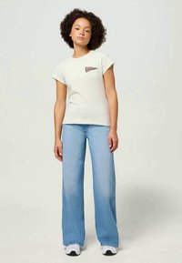 Cremfarbendes Kurzarm-T-Shirt mit einem Logo-Patch, kombiniert mit hellblauen weit geschnittenen Jeans und weißen Sneakers. Glatter Stoff und lässige Passform.